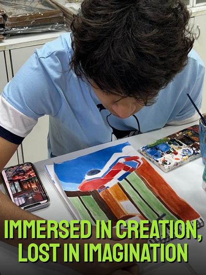 Immersed in creation, lost in imagination. A young student is deeply focused on his painting, using a reference photo on his phone to capture the details perfectly.