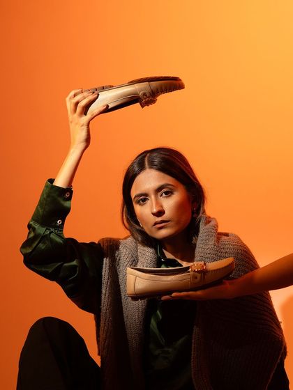 A creative, editorial-style fashion shoot featuring loafers. The model's pose and the bold orange background create a high-fashion, confident look.