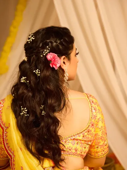This half-up, half-down hairstyle for a Haldi ceremony is both beautiful and practical. It features a braided crown and soft waves, decorated with baby's breath and a single bright flower for a pop of color.