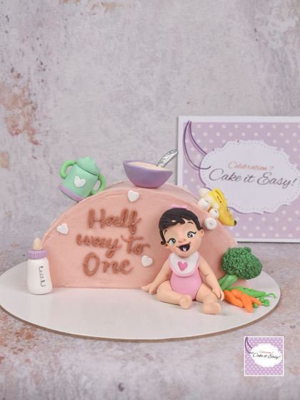 A unique cake to celebrate a baby being 'Halfway to One'. This top-forward cake tells the story of starting solid foods, with fondant milestones from a milk bottle and sippy cup to a bowl of porridge and fresh vegetables.
