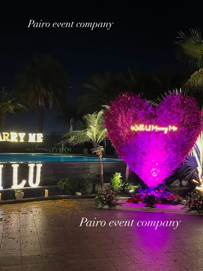 A complete proposal scene by the pool, featuring a large purple-lit floral heart, "I L U" and "MARRY ME" marquee letters, creating a spectacular and romantic setting.
