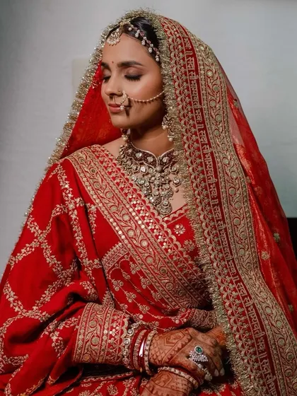 A close-up of a traditional bride in a heavily embroidered red velvet outfit. The makeup features soft, defined eyes and a flawless base to balance the richness of her attire.