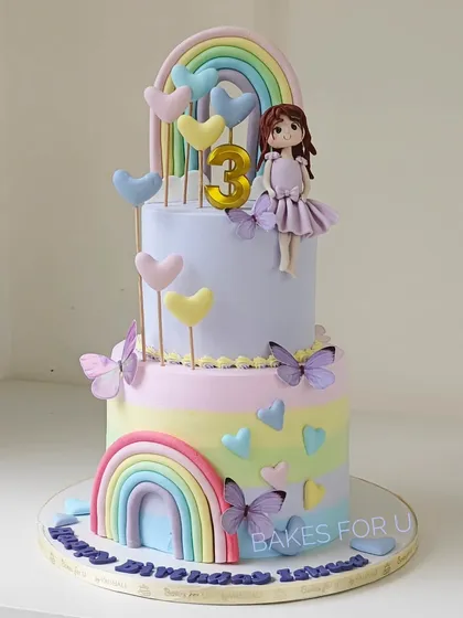 A sweet and colorful two-tier cake featuring rainbows, butterflies, and a cute fondant girl topper. The pastel palette and heart accents make this a lovely choice for a young child's birthday.