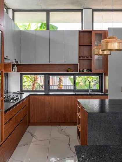 This modern kitchen combines teak wood cabinetry with grey upper units and a black granite countertop. High clerestory windows bring in ample natural light, reducing the need for artificial lighting during the day.