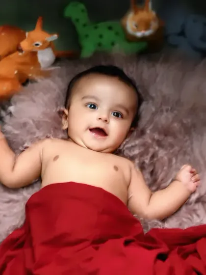 A happy baby lies on a soft fur rug, surrounded by greenery and woodland animal toys, smiling up at the camera.