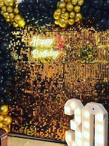 A gold shimmer wall paired with black and gold balloons creates a very glamorous and high-energy photo corner. This setup for a 30th birthday features a 'Happy Birthday' neon sign and light-up numbers, perfect for a stylish and modern celebration.