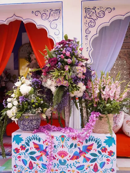 A beautiful console table setup, featuring a hand-painted floral cloth and large bouquets of purple and white flowers. The backdrop of colorful drapes in traditional arches adds to the royal Rajasthani vibe.