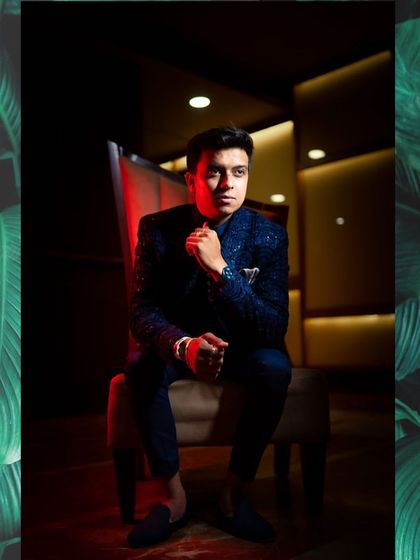 A stylish portrait of a groom in a modern blue suit, seated in a chair with dramatic red and dark lighting creating a moody and sophisticated look.