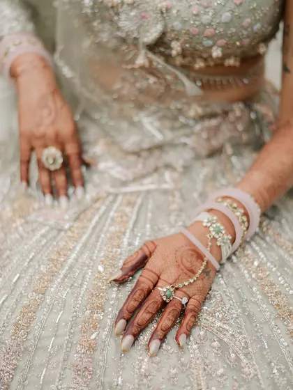 The delicate details of a bridal look. This close-up showcases the intricate henna, the modern hand jewelry, and the soft pastel tones of the bride's heavily embroidered lehenga, all part of her curated style.