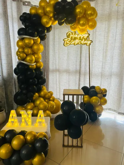 A simple and chic black and gold birthday setup. The balloon half-arch and 'Happy Birthday' neon sign create an elegant photo corner for a sophisticated celebration.