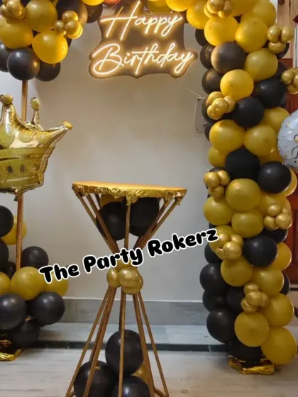 A stylish and modern birthday setup in black and gold. The decor includes a balloon arch, a neon sign, a diamond-shaped cake stand, and a crown foil balloon pillar.