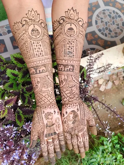 A full bridal mehendi design that goes up to the elbows, showcasing traditional Indian artistry. The palms feature classic dulha-dulhan (bride and groom) figures, while the arms are filled with intricate patterns, including elephants, kalash motifs, and delicate jaal work, all done with natural henna.