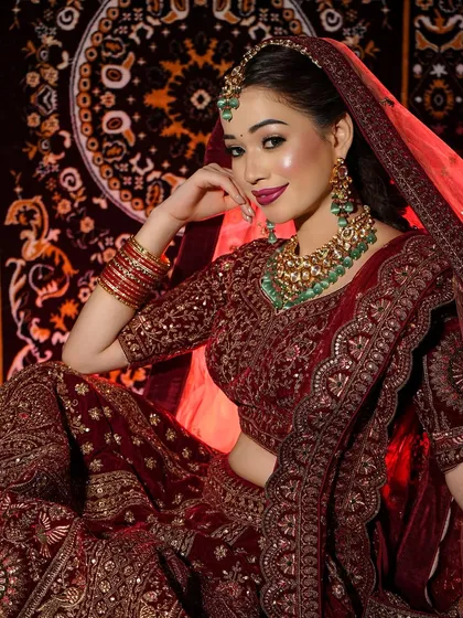 A retro-inspired bridal look with a deep red velvet lehenga, bold lips, and winged eyeliner, complemented by green kundan jewelry.