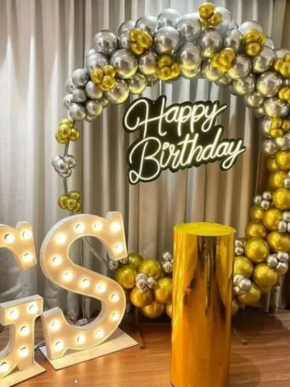 A glamorous gold and silver themed party. This setup includes a circular balloon arch, a neon sign, a reflective gold pedestal, and large light-up marquee letters for initials.