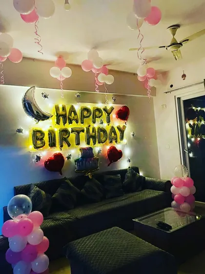 A cozy living room setup for a birthday surprise. This decoration includes 'Happy Birthday' foil balloons, fairy lights, and pink and white balloons on the ceiling and floor.