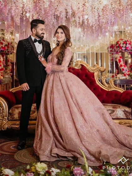 A beautiful couple posing for a photo at their reception. The bride is wearing my elegant dusty pink long-sleeved gown with feather cuffs and a full, sparkling skirt.