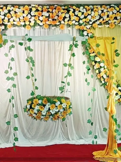 An elegant naming ceremony setup with a hanging cradle. The stage is decorated with yellow and white flowers, flowing drapes, and traditional greenery.