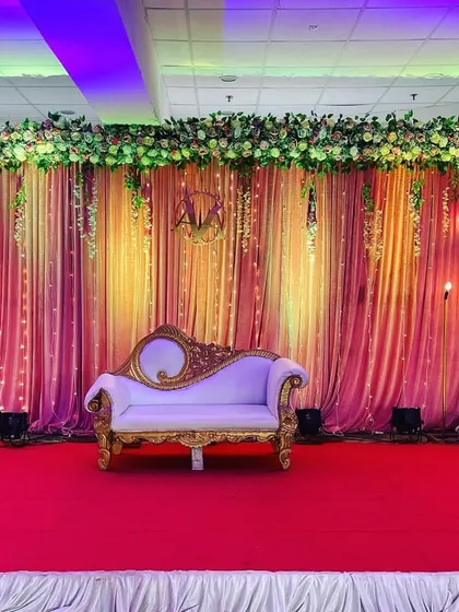 A classic reception stage with a golden-pink drape backdrop, hanging floral arrangements, and a vintage-style sofa for the couple.