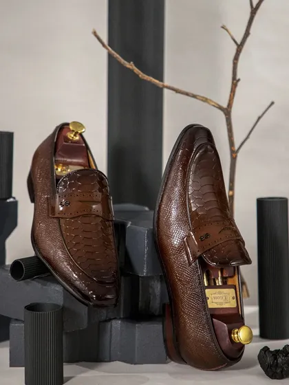A styled shot of men's textured leather loafers. The use of dark, geometric props and a single branch creates an artistic composition that feels both modern and timeless.