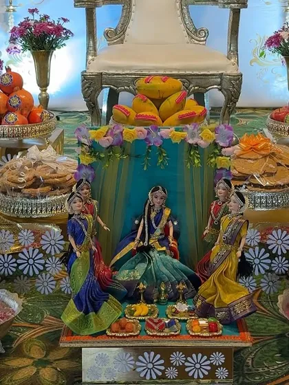 A close-up of a beautiful doll arrangement depicting a Seemantha ceremony. The dolls are surrounded by offering plates filled with traditional sweets and snacks.