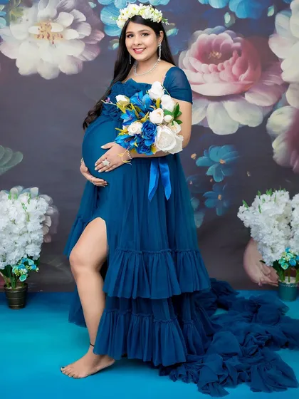 A vibrant and joyful solo portrait celebrating the final stages of pregnancy. The mom-to-be glows in a royal blue ruffled gown, complemented by a lush floral backdrop and a matching bouquet, creating a rich, colorful scene.
