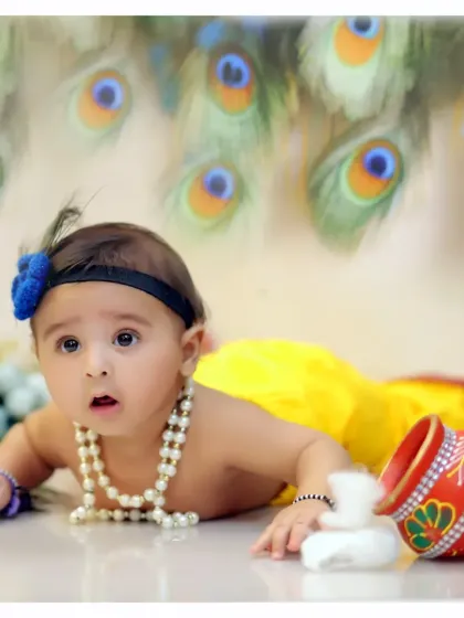 A wide-eyed baby lies on their tummy during the Krishna photoshoot, surrounded by props like a painted matka. The peacock feather background adds a touch of divine charm.