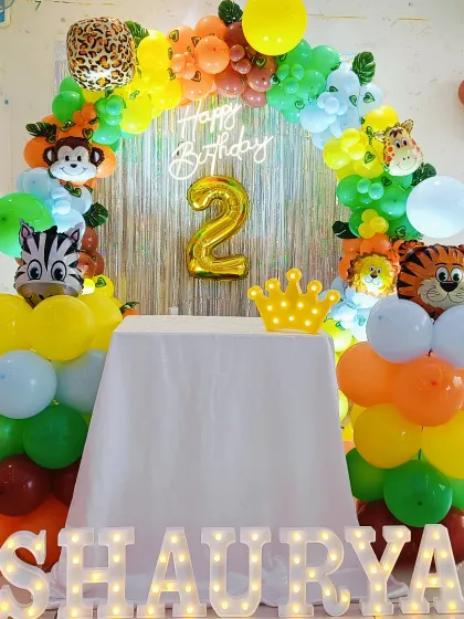 A fun jungle-themed second birthday party. The setup includes a colorful balloon arch with foil animal heads and a 'Happy Birthday' neon sign against a tinsel curtain.