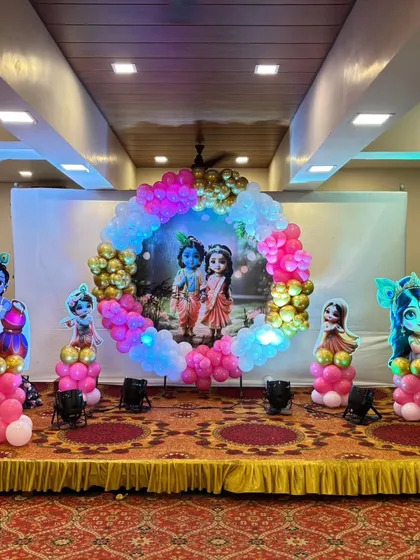 This Radha Krishna themed baby shower stage features a large circular balloon arrangement around a central backdrop, with character cutouts on either side.