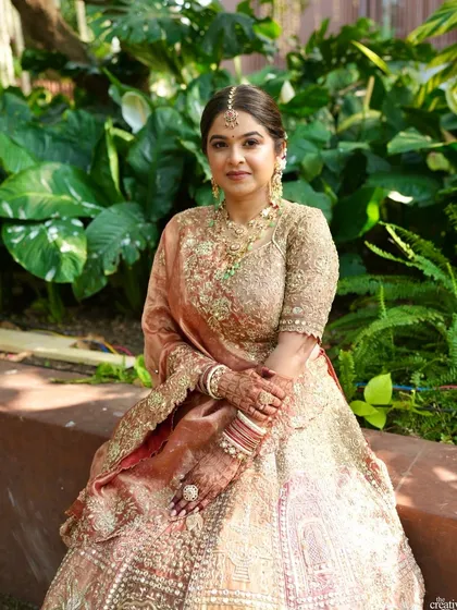 A full portrait of the bride seated against lush greenery. The pastel colors of her lehenga and the soft, natural makeup look absolutely ethereal.