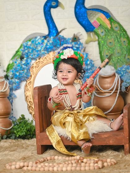 Hare Krishna! This little one is sitting happily on a small wooden bench, holding a flute, in a beautiful peacock-themed setting.
