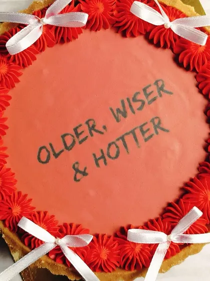 Celebrate with a bit of sass! This chocolate tart features the fun message 'Older, Wiser & Hotter', perfect for a birthday with personality. The bold red design is finished with delicate white ribbons for a playful touch.