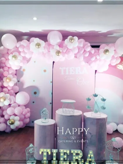 A pretty-in-pink first birthday party with a daisy motif. The setup features a large pink balloon arch with daisy accents, a custom backdrop, and themed cake stands.