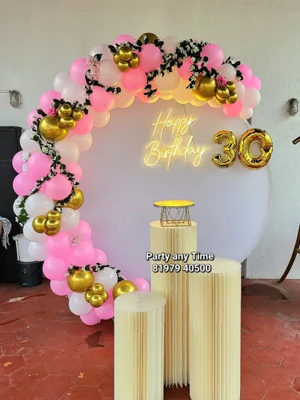 A chic 30th birthday celebration with a pink, white, and gold balloon arch. The setup includes a '30' foil balloon and a "Happy Birthday" neon sign.