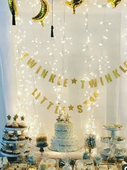 The 'Twinkle Twinkle Little Star' theme set up for a baby shower dessert table. This decoration features a curtain of warm fairy lights, a golden banner, and foil moon and star balloons, making the cake and treats stand out.