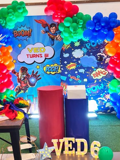 A powerful superhero-themed party for Ved's 6th birthday. This setup includes a comic-book style backdrop with Superman and Iron Man, themed plinths, and marquee letters.