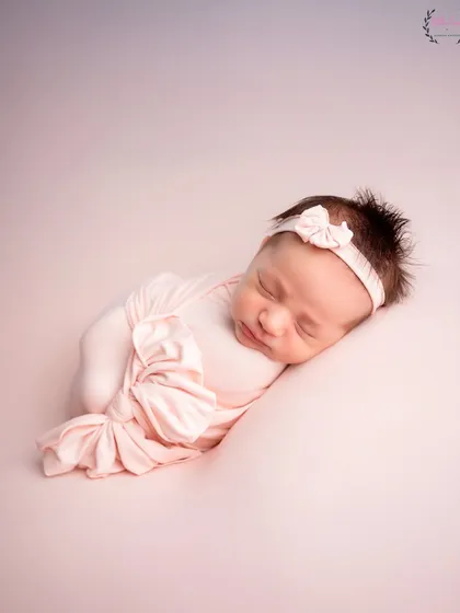 A newborn baby is swaddled in a soft pink wrap with a matching bow headband, sleeping on a pink surface. This photo is a perfect example of a minimalist and color-themed approach, focusing entirely on the baby.
