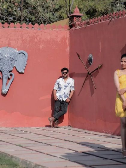 A stylish and moody pre-wedding shot with an interesting composition. The bride is in the foreground while the groom leans against a vibrant red wall, creating a cool, editorial feel.
