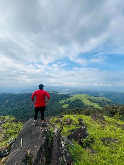 A trekker stands in a power pose on a summit, looking out at the rolling hills of the Western Ghats.