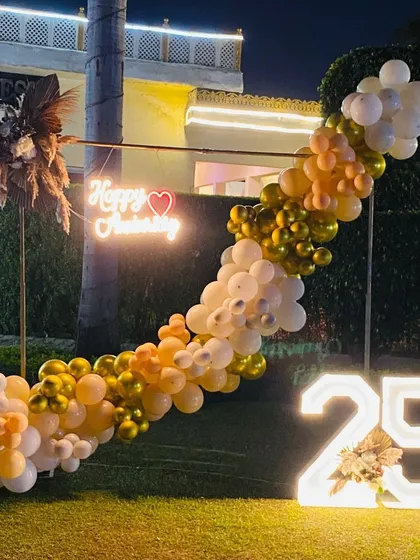 Celebrating a silver jubilee anniversary outdoors. This elegant setup features a custom 'Happy Anniversary' neon sign, a flowing balloon garland in gold and white, and a large light-up '25' to mark the special milestone.