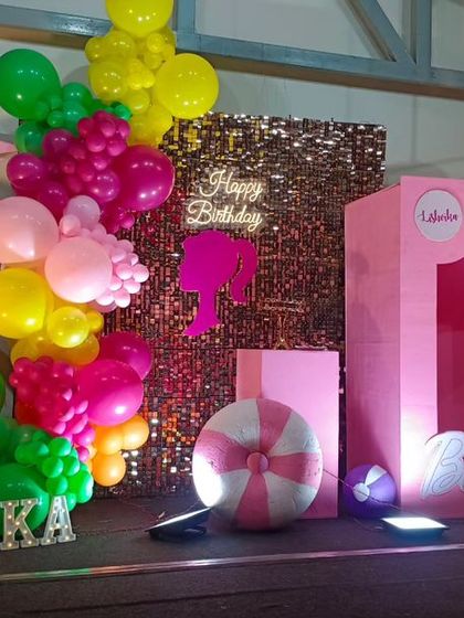 A vibrant, tropical Barbie-themed birthday party. This setup features a pink "doll box" photo booth, a sequin backdrop, and colorful balloon clusters with palm tree cutouts.