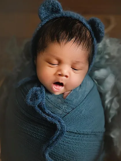 A close-up of a 10-day-old newborn yawning while wrapped in a cozy blue swaddle and bear-ear bonnet. This candid capture shows the beauty in the unposed, natural moments of a newborn session.