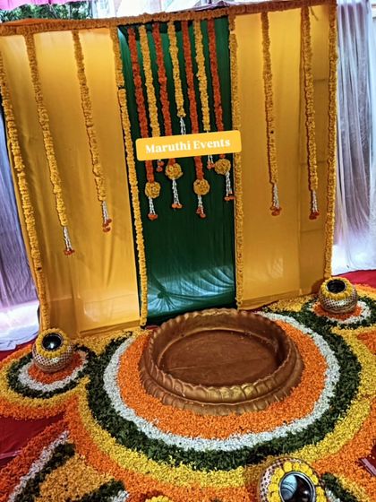 A traditional setup for a havan or puja, featuring a backdrop of yellow and green drapes with marigold garlands. The floor is covered with a floral rangoli, creating a sacred space for rituals.