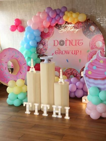 A fun 'Donut Grow Up' themed birthday party. The setup includes a donut-print backdrop, large donut and cupcake props, and a colorful balloon garland.