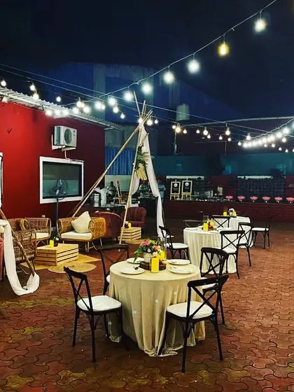 A rustic and inviting outdoor event setup. The space features bohemian-style lounge seating with teepees, round dining tables, and a buffet station in the background, all under a canopy of warm string lights.