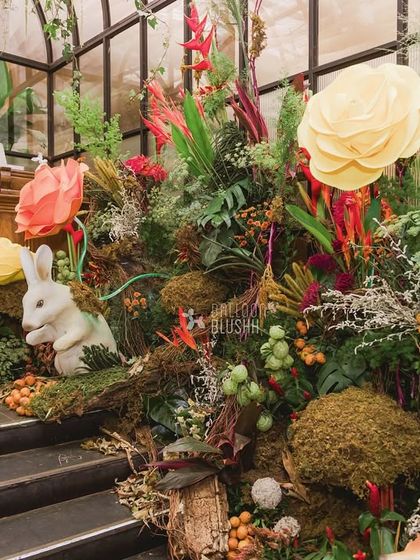 A white rabbit figurine sits among a lush arrangement of moss, branches, and colorful flowers on the stairs.