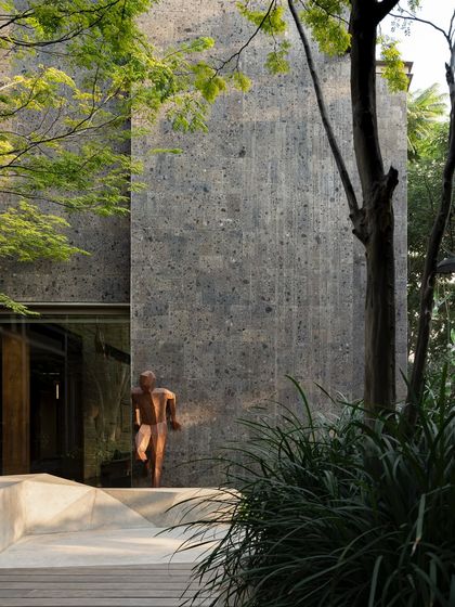A bronze sculpture is strategically placed at the entrance of a building, framed by trees and rough stone walls. The artwork guides the eye and adds a touch of classical elegance to the modern landscape.