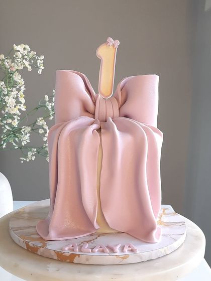 Tying up the cutest first birthday in a bow. This elegant and simple cake is wrapped in a large, realistic, and fully edible pink bow, making it look like the perfect birthday present.