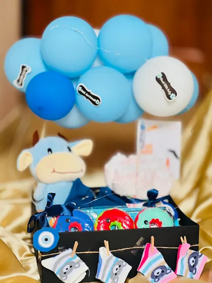 Welcoming a baby boy with this essentials hamper. It includes a soft cow plush toy, colorful rattles, diapers, and adorable printed bibs, all arranged in a handsome black crate decorated with a 'Just Arrived' balloon bouquet.