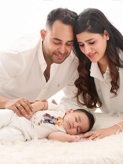 The whole family, dressed in white, gathers around their sleeping baby, creating a peaceful and loving scene on a soft, white fur rug.