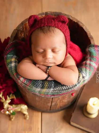 A close-up of a newborn nestled in a wooden bucket, wearing an adorable red bear bonnet. The warm wood tones and soft textures create a cozy, rustic feel.